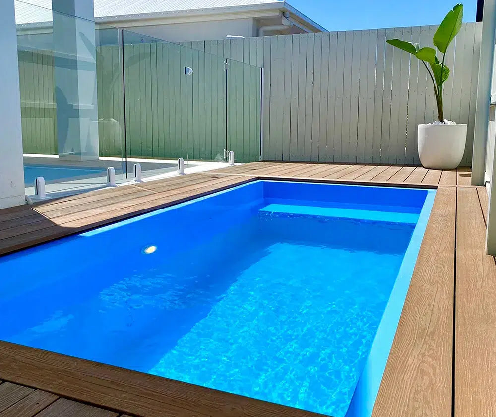 Modern fibreglass pool with wooden deck and glass fence on a sunny day